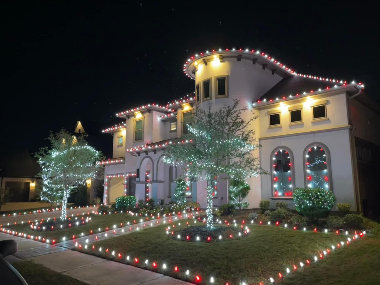 Two-story Sienna home with red and white C9 roofline lights, wrapped trees and lit lawn borders